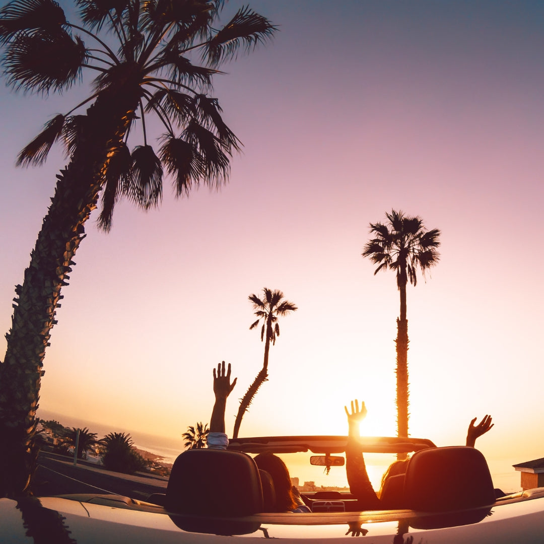 Zwei Personen fahren in einem Cabrio unter Palmen in den Sonnenuntergang, mit erhobenen Armen und einem Gefühl von Freiheit.