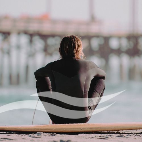 Ein Mann im schwarzen Neoprenanzug sitzt auf einem Surfbrett am Strand, schaut in Richtung Meer und Pier. Über dem Bild liegt ein weißes Symbol mit zwei geschwungenen Wellenlinien.