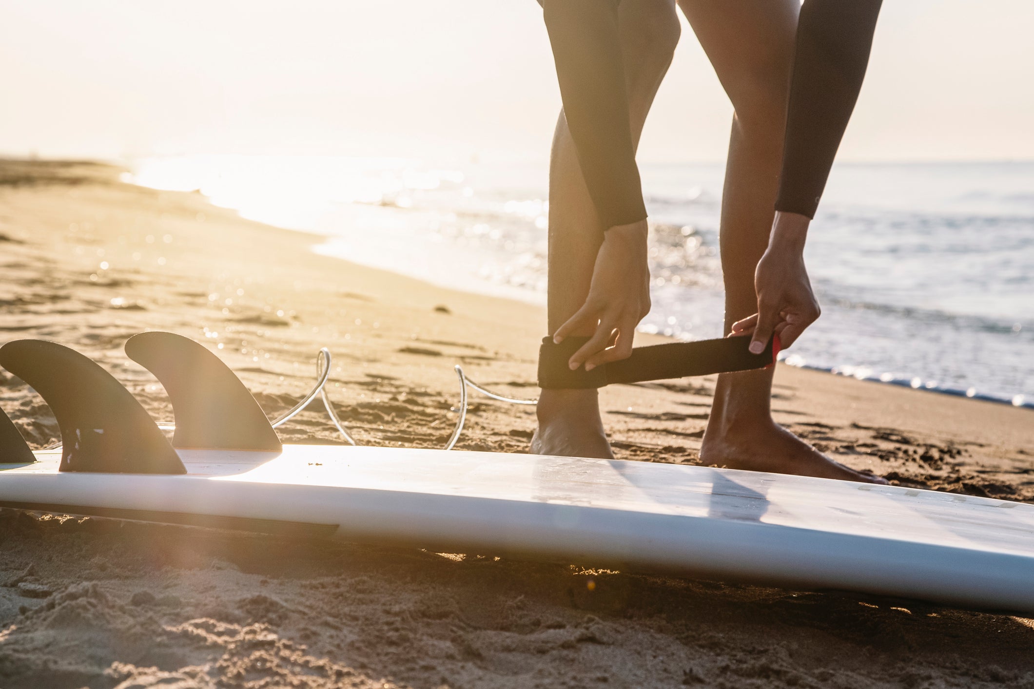 Surfer am Strand im Morgenlicht, der ein schwarzes Surfboard Leash anlegt. Im Vordergrund liegt ein Surfbrett mit Finnen im Sand, das Meer glitzert im Hintergrund in der aufgehenden Sonne.